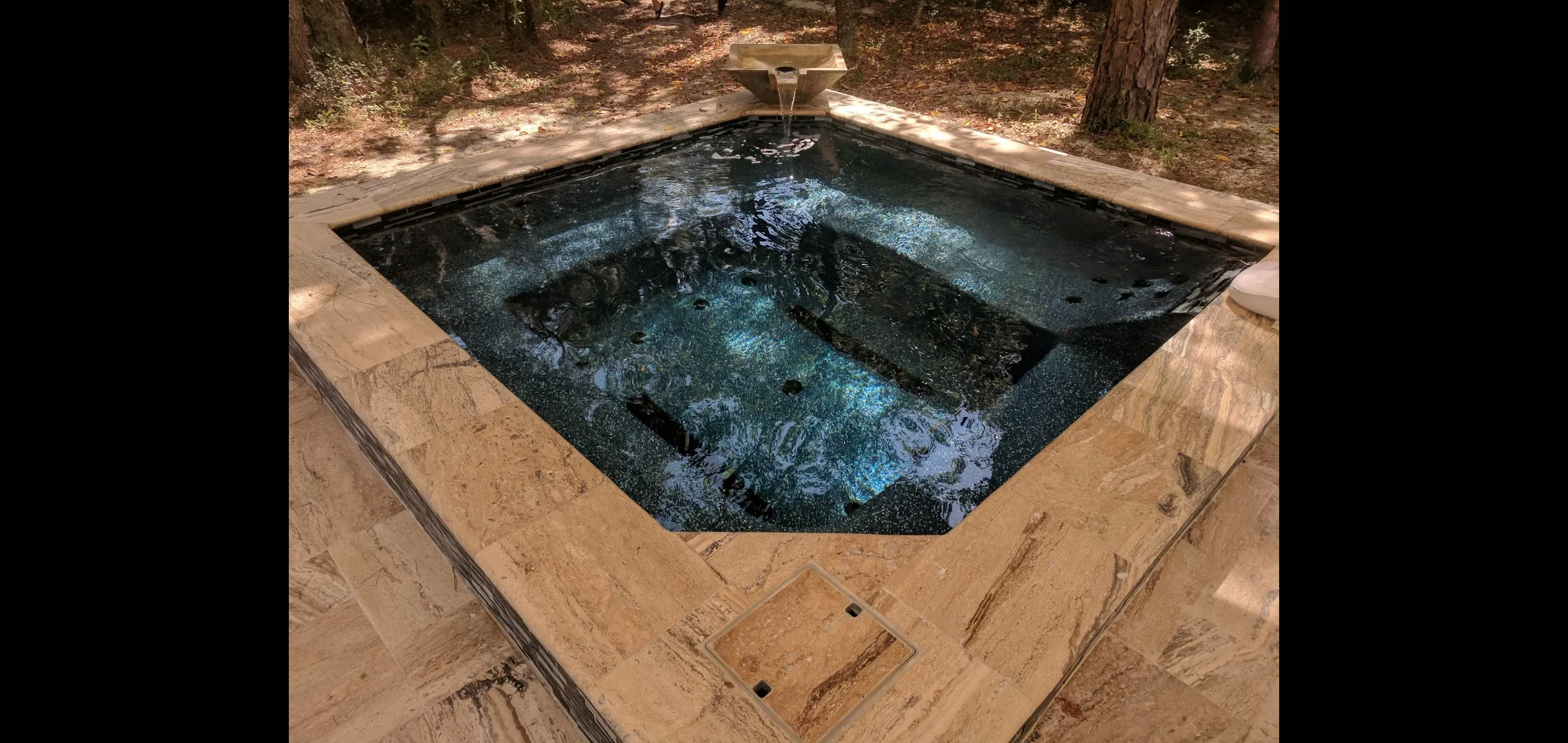 In-ground spa with dark aggregate finish, travertine deck, and waterfall feature in wooded setting